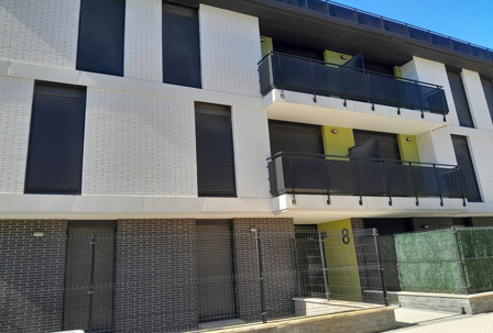 Fachada de edificio residencial de varias plantas con balcones, persianas oscuras y acceso vallado en planta baja.